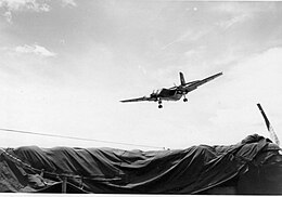 Large airplane descending over tarpaulin-covered area