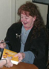 A woman sitting a table with the shot focusing on her upper body. She is black and blue clothing.