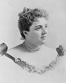 A young white woman with curly dark hair in an updo, wearing a gown with bare shoulders, and pearls