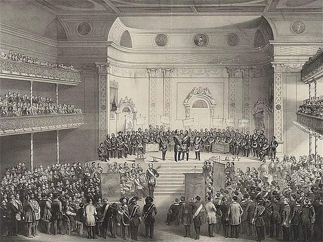 Drawing of interior of large concert hall, crowded with formally dressed people, mostly men. There are two balconies. The main floor has no seats, so attendees are standing. The stage is crowded with male participants in a formal ceremony.