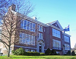 A two-story brick building seen from its left, with a bare tree in front on that corner. It has many large windows, two entrances with decorative stone trim and a point in the center of the roof. The U.S. and POW-MIA flags are flying from a flagpole in front