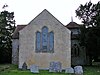 The end of a stone-built church, with a triple window overlooking some gravestones. Flint-built walls and a small belfry are visible behind.