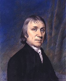 Quarter-length portrait of a man in a black coat against a purple and blue curtain backdrop.