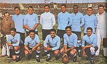 Photo d'avant match d'une équipe de football habillée de bleu, accompagnée de deux officiels, au milieu d'un grand stade.