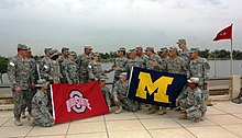 Two groups of soldiers in camouflage uniforms staring at each other on a waterfront; the left group carries a red flag with a silver "O" and the words "OHIO STATE" on it; the right group carries a blue flag with a yellow "M" on it.