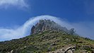 Das Foto zeigt einen Berg, auf dessen Gipfel hohe, zusammenhängende Säulen aus Phonolithgestein stehen. Darüber wölbt sich bogenförmig eine Wolke.