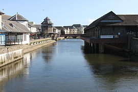 La rivière canalisée et entourée de bâtiments, dont un marché à droite et une tour à horloge à gauche.