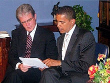 A man with glasses and Obama sit and hold a sheet of paper. Obama points at the paper and talks. Both men wear dark suits and ties.