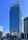 Ground-level view of a blue, glass high-rise; the tower sits behind a small, white, stone, window-dotted facade