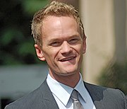 An image of a blonde-haired Caucasian male in his starting 40s smiling. He is wearing a grey-striped blazer with a white dress shirt and a silver tie.