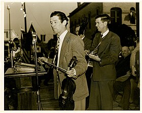 un homme en veste debout sur une scène devant un micro, avec un violon dans une main et un archet dans l'autre, musiciens derrière lui