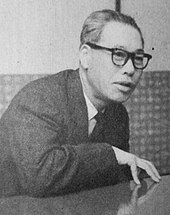 Publicity still of Shimura cleanly shaven and wearing glasses.
