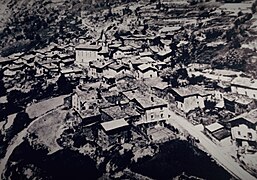 Photographie aérienne sur une carte postale en noir et blanc : le village est circulaire, des maisons par dizaines entourent l'église au centre, il y a plusieurs chapelles et la route dessert des ruelles.