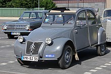 Ein grauer Citroën 2CV Baujahr 1956 steht auf einem Parkplatz, hinter ihm zwei weitere Fahrzeuge. Das Fahrzeug entspricht den typischen 2CV seiner Bauart. Er steht frontal zum Betrachter, das Faltdach ist geöffnet und am linken Kotflügel befindet sich ein blauer Aufkleber mit der Aufschrift „Moselle“.