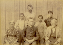 Formal photograph of an older Eliza and Mathias Splitlog, seated with a young priest wearing a large crucifix. Behind them are two Splitlogs of the younger generation, and two grandsons.