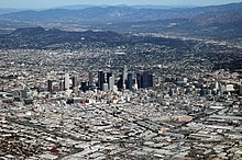 An aerial view of a widespread built-up area, skyscrapers in the central district, with mountains in the background.