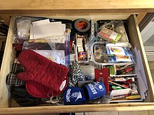 A junk drawer in a residential kitchen. As is typical of junk drawers, this drawer contains various unrelated and unorganized items including beer can koozies, birthday candles, matches, hand sanitizer, paper clips, oven mitts, black electrical tape, a yellow screwdriver, pens, pencils, a green manual pencil sharpener, plastic bags, rubber bands, loose slips of plain white notepaper, and white envelopes.
