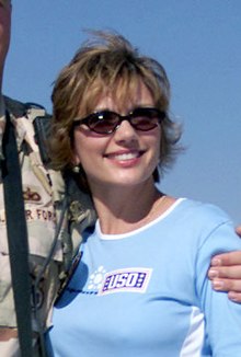A candid photo of a white woman in sunglasses and a "USO" shirt; an arm is wrapped around her back, holding her left shoulder, and she is looking to the camera's right.