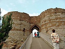 Two round towers of light yellow stone at the bottom and dark orangy stone at the top on either side of an arched entrance. A bridge leads from the entrance to allow access.