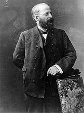 Eduard Bernstein standing next to a chair and looking rightwards. He is resting his hand on the chair.