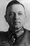 The head of a man, shown in semi-profile. He wears a military uniform with a military decoration in shape of an iron cross displayed at the front of his shirt collar. His hair is dark e combed to back.