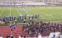 A high school football field with a large "DV" emblem in the center of the turf. Dougherty Valley football players in blue and silver uniforms are lined up on the side of the field. On the track surrounding the field, there are cheerleaders in light blue, white, and navy blue uniforms with blue pom-poms to the left. To the right of the track there are members of the pep band with their instruments in black shirts.