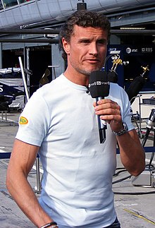 Photo d'un homme en tee-shirt blanc, un micro à la main, devant des stands de course