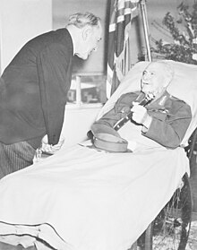 A man lies in a hospital bed, incongruously wearing an Army uniform instead of pyjamas. His peaked cap is on the blanket and he holds a baton in his hand. A man in a dark suit and pinstripe trousers bends over to talk to him. In the background are flowers, and a flag.