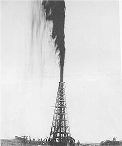 Black-and-white photograph of an oil derrick with a gusher of oil shooting from the top