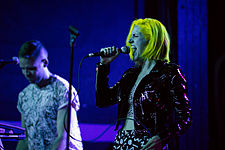 A woman with yellow hair sings into a microphone on stage, with a man playing the keyboard to her left.