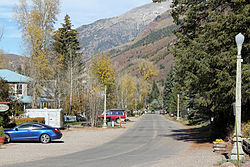 A street lined with streetlights and houses on large lots sheltered by trees, leading towards tall mountain slopes in the distance