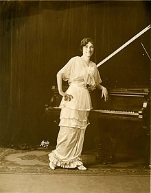 A slim young white woman stands leaning one elbo on a piano, hand on hip; she is wearing a floor-length tiered light-colored gown