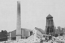 Photo en noir et blanc montrant des hommes sur des pierres, à l'arrière-plan une cheminée et une tour en bois (chevalement).