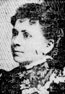 A middle-aged white woman, wearing a high-collared lacy dress or blouse, and an updo with curls across her forehead