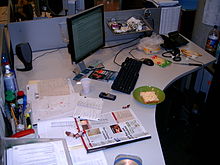 Una scrivania da ufficio in un cubicolo, che mostra la condivisione dello spazio tra componenti del computer e documenti cartacei