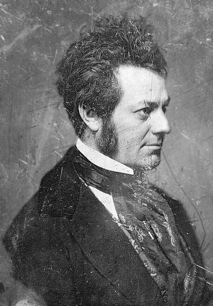 Scratched black and white photo of Edwin Forrest in profile intently looking out ahead. His dark hair is longer on top and slightly wild. He wears mutton-chop facial hair. He wears a dark jacket, starched white color, and voluminous dark patterned necktie that cascades down his breast. 