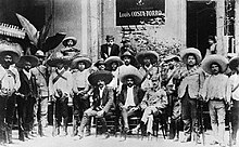 Emiliano Zapata sits in a chair surrounded by his supporters and other Zapatistas.