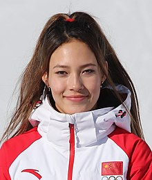 Young smiling woman with brown hair pulled back in a ponytail, wearing earrings and a red-and-white ski jacket with the Chinese and Olympic flags. She is standing in front on a white background.