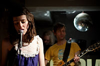 A brunette woman singing into a microphone. In the background there is a male guitarist wearing a yellow t-shirt, and a glitter ball