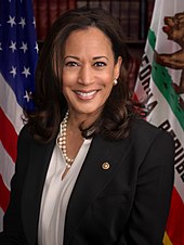 A woman in front of the U.S. and California flags, wearing a black suit and multiple pearl necklaces, and smiling in front of the camera