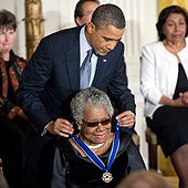 An elderly African-American woman, seated, smiling broadly, and dressed in black, being given an award by an African-American man in his fifties, wearing a blue tie and leaning over from behind her.