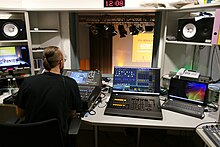 Ein Mann mit Pferdeschwanz ist von hinten zu sehen, er sitzt vor einem Mischpult in einem Raum mit weiteren technischen Geräten und mehreren Monitoren.