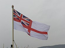 A flag waving in the wind. The flag is on a ship. The flag is white and has a red cross on it. There is a British flag in the top left of the flag.