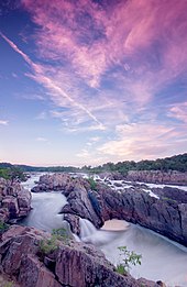 Rapids in a wide, rocky river under blue sky with clouds colored purple by the sunset.