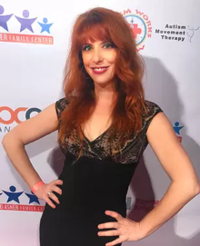 A woman with long, red hair stands in a black cocktail dress facing the camera. Behind her is a red-carpet background for the Ed Asner Family Center.