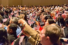 Dozens of adults sit in auditorium rows, many waving small American flags