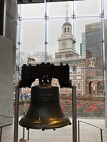 The Liberty Bell hangs in a glass-backed structure with a brick, 18th-century building with a steeple visible in the background.
