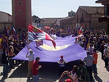 People at a celebration holding a huge purple flag.  Others wave different flags, such as the Cross of Burgundy or the modern flag of Castile and Leon.