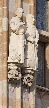 Zwei helle Steinfiguren auf Konsolen an einem Renaissance-Gebäude. Links im Gewand eines Mönchs mit einem Lesepult, rechts ein Ritter mit Schild und Schwert Ritter