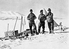 Photographie noir et blanc de trois hommes en tenue polaire, une tente derrière eux et un traineau à leur droite.